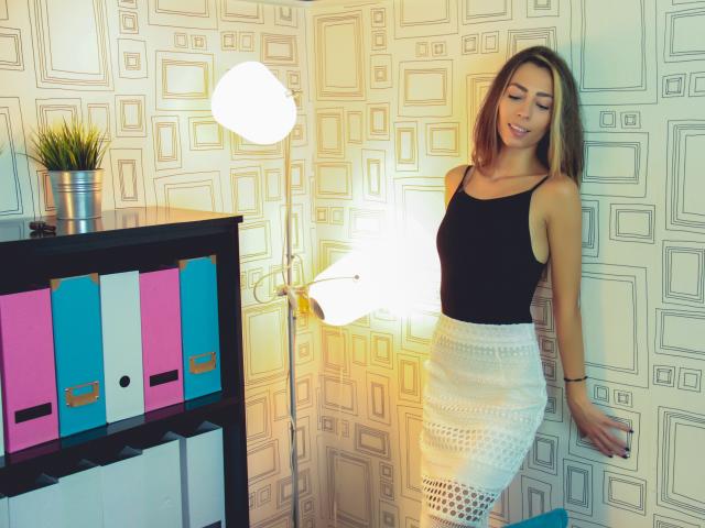 PoxyVibe with medium tits, wearing a black tank top and white lace skirt, stands by a lit floor lamp, next to a black shelf with pink and blue binders.