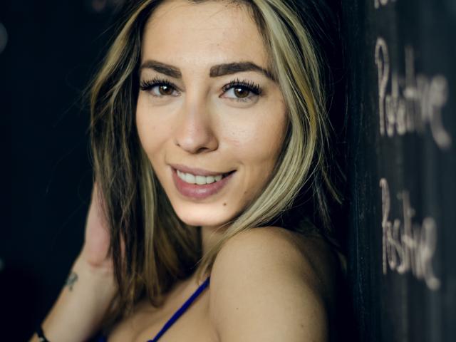 PoxyVibe has blonde and brown hair, brown eyes, and is smiling with a cute face, showing her tits in a blue top, with a small tattoo on her arm.