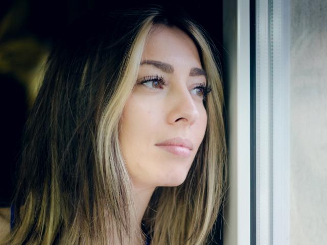 PoxyVibe's face, showing a slight smile, with blonde and brown hair, blue eyes, and long eyelashes, standing near a window, looking outside. Her tits are not visible.