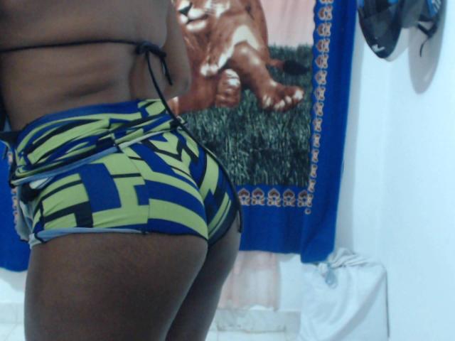 SamyHotCool's dark brown butt in tight, blue and green striped shorts, back to the camera, wearing a black string bikini top, in a brightly lit room.