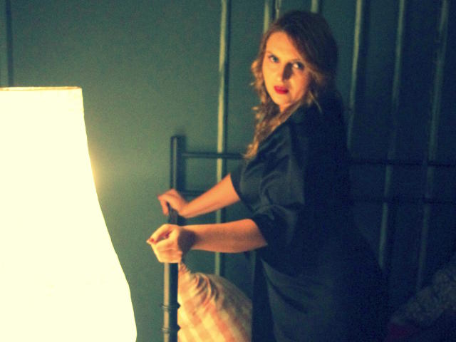 Oliviaaa with wavy brown hair, red lipstick, and black dress, leaning on bedpost, holding it with hands, standing next to bright lamp, in dimly lit room.