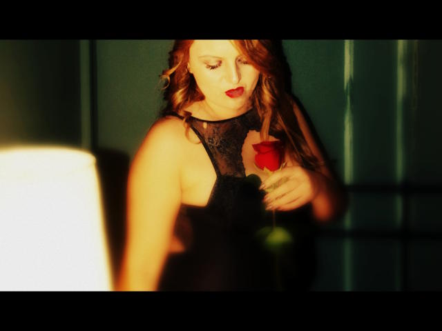 Oliviaaa, avec des cheveux roux, portant une robe noire en dentelle, tenant une rose rouge, regardant vers le bas, éclairée par le soleil venant de la gauche, fond vert foncé, lumière douce d'une lampe sur la gauche..