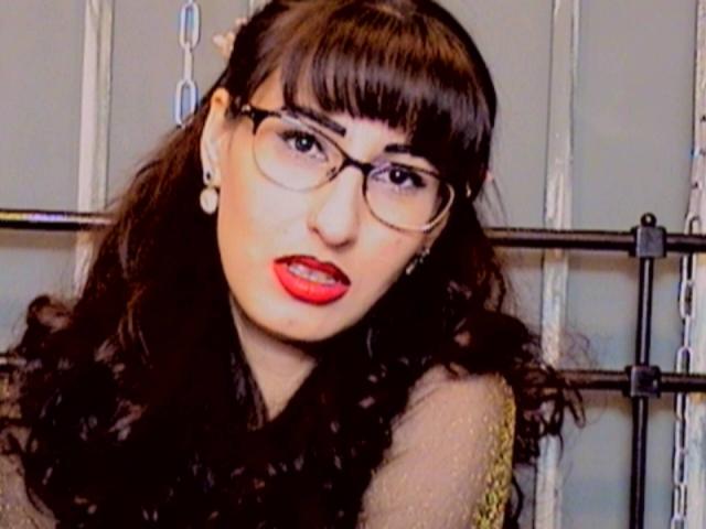 LysAddiction with dark brown hair, bangs, glasses, red lipstick, and a sheer top, showing off her tits, looking sexy, standing in front of a metal rack.