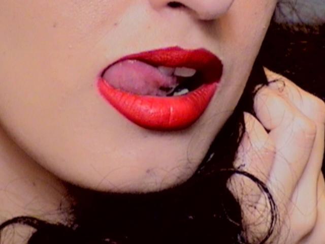 LysAddiction's red lips are slightly open, showing a small bit of pink tongue. Her hand is near her face with dark, wavy hair visible. Her skin is light.