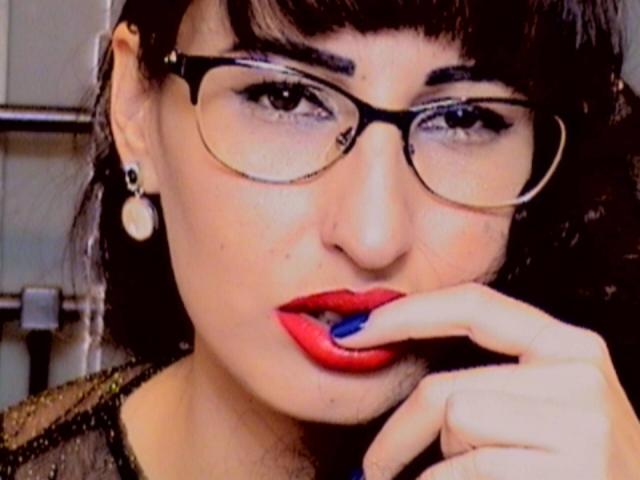 LysAddiction has red lipstick, blue nail polish, and is sucking her finger with glasses on, black hair, and a black sheer top. She's got a nose piercing and ear studs.