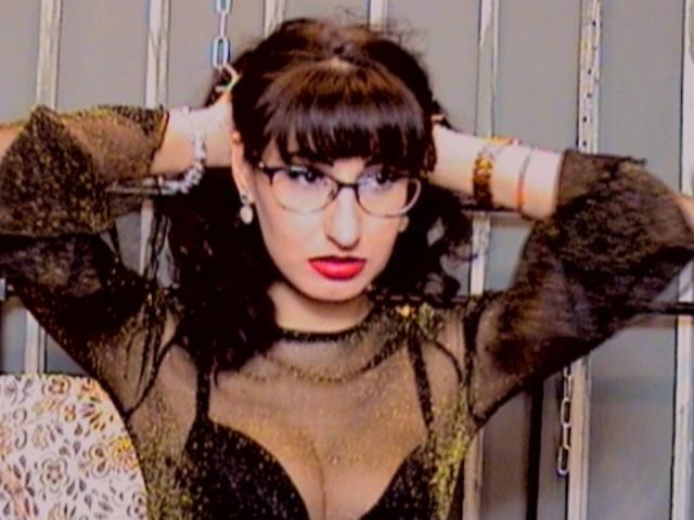 LysAddiction with glasses, black lace top, red lipstick, arms raised, hands in hair, showing her big tits, black hair, wearing jewelry, against a metal wall.