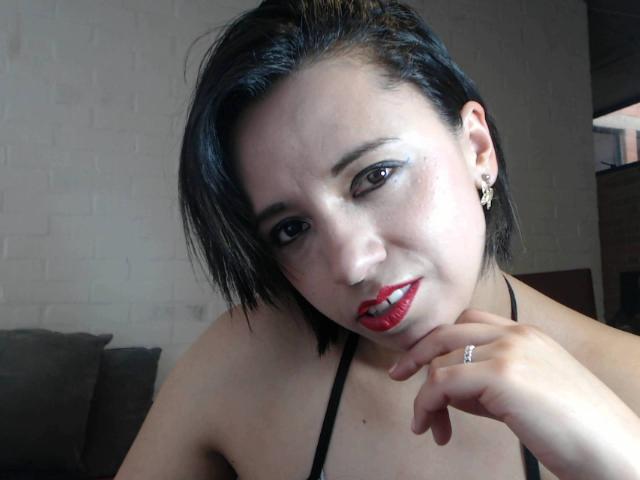 BellaLolita with short black hair, red lipstick, and black eyeliner, smiling at the camera, hand near her chin, wearing a black strap, showing her small tits and ring.