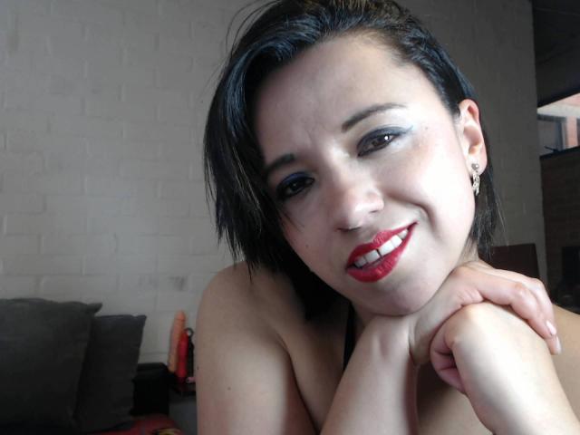 BellaLolita with short black hair, red lipstick, and blue eyeshadow, smiling at the camera, hands resting on her knees, topless, showing her ass, in a cozy room.