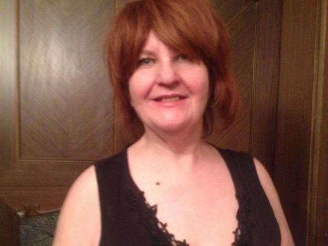 MioritaStar with short red hair, smiling, wearing a black lace top, showing her boobs, fair skin, small mole on her chest, wooden door background, casual selfie.