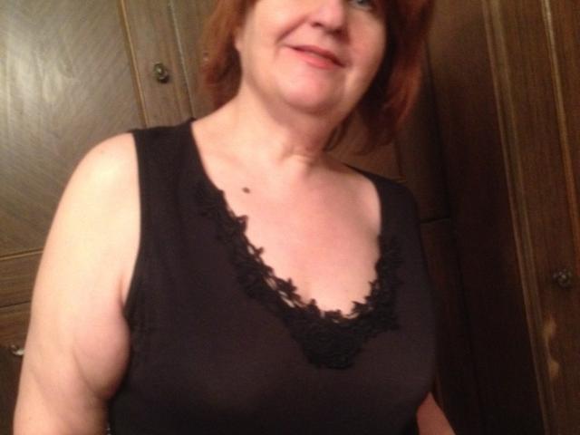 MioritaStar, middle-aged woman with red hair, smiling, wearing a black lace-trimmed sleeveless top, showing her tits, wood cabinets in background, fair skin, small mole on her chest.