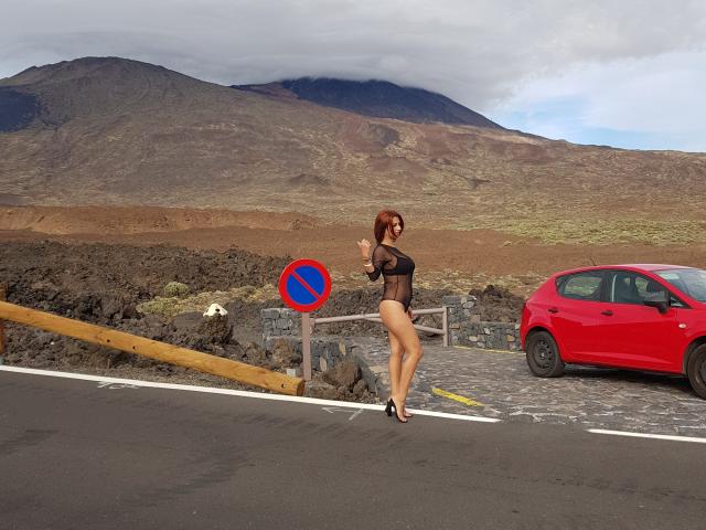 IssaDiamondd, in black sheer lingerie, standing on a road with a red car, making a peace sign, has red hair, small tits, and a curvy ass, wearing black heels.