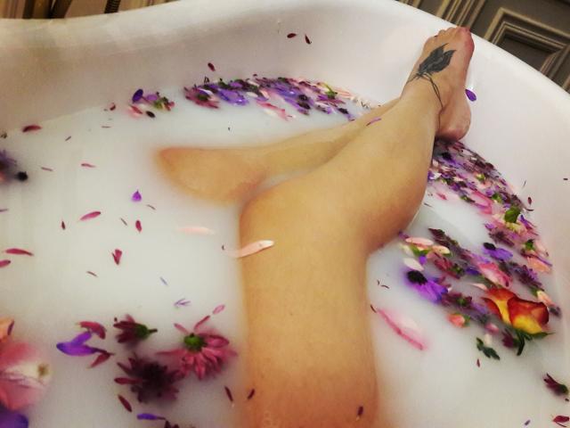 ValeriMair's legs in a white bathtub filled with water and colorful flower petals, with a black tattoo on her right ankle, showing a relaxed and sensual pose.