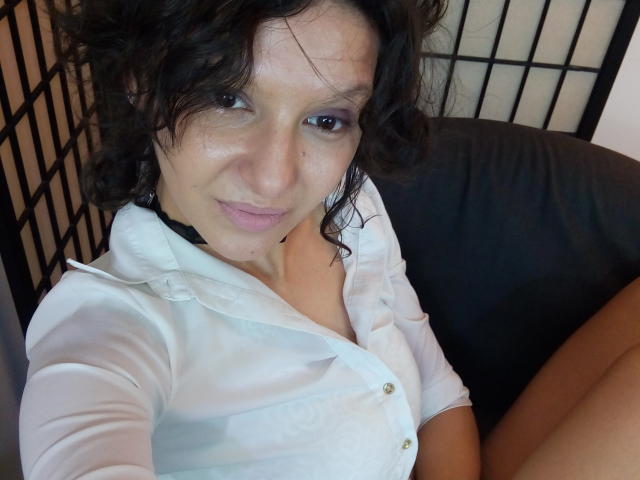 AmethystCharm has curly brown hair, light skin, and wears a white button-up shirt with a black choker, sitting on a black couch, showing a slight smile and relaxed expression.