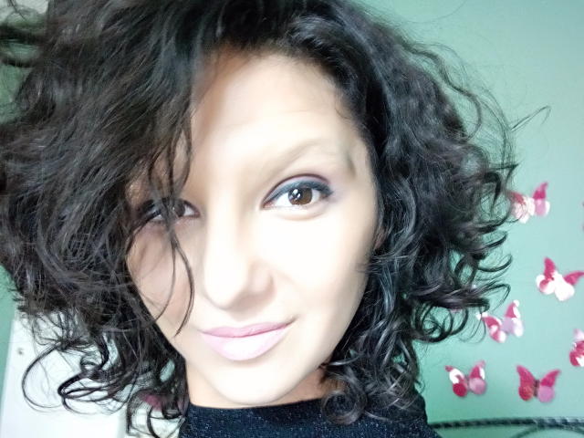 AmethystCharm has curly dark hair, brown eyes, and pink lipstick, smiling slightly with a subtle nose. She's wearing a black top with pink butterfly stickers in the background.