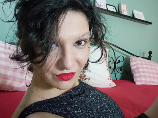 AmethystCharm has dark, wavy hair, red lipstick, and a sparkly black top, looking up with brown eyes. She's on a red bed with pink checkered pillows.