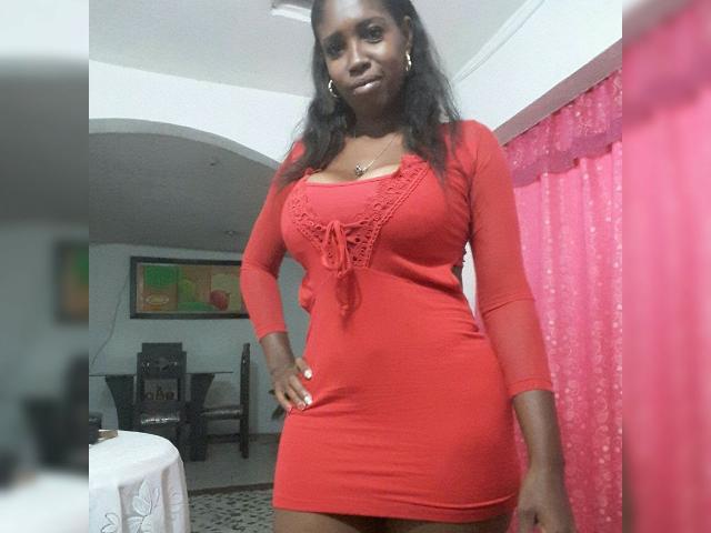 AgataFoxx in a tight red dress with long sleeves, showing her tits and ass, standing with one hand on her hip, looking at the camera, in a room.