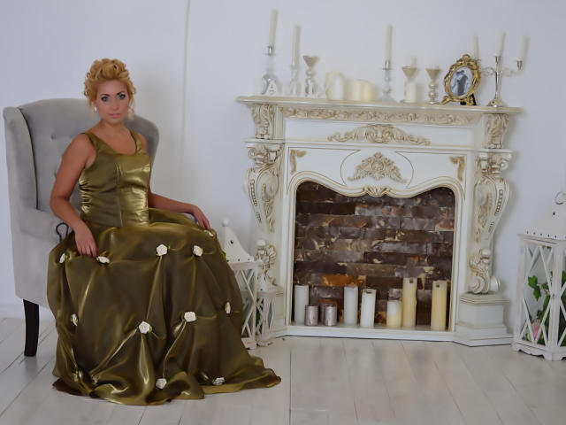 IrickaKicka with golden satin dress, white flowers, sitting on gray chair, arms on knees, next to ornate white fireplace, candles, and white lanterns, blonde hair, blue eyes.