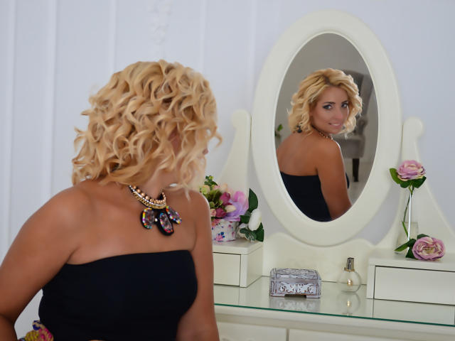 MarySunny with curly blonde hair, black strapless top, and colorful necklace, looking at herself in an oval mirror. She has tan skin and a cute ass reflected.
