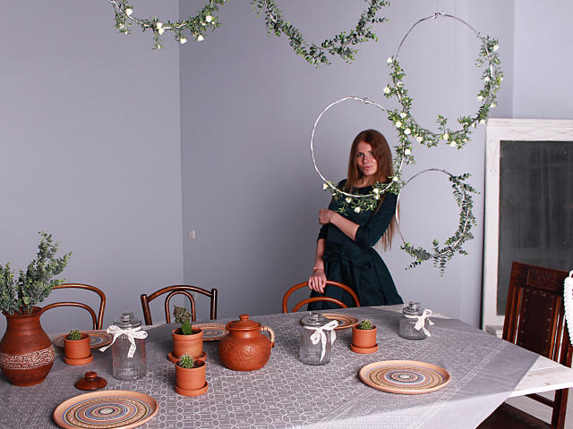 GretyArdent with long brown hair, wearing a dark green dress, stands by a table with terracotta pots, white ribbons, and green plant wreaths. Her tits are small, she's cute.