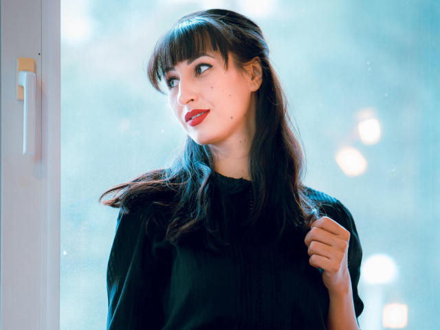 LysAddiction with dark brown hair, red lipstick, and black shirt, holding shirt with right hand, looking to the side, window in background, soft blue light, slight smile.