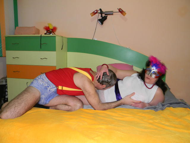 A personagem Keyx, com uma máscara rosa e azul com penas, uma camisa branca e shorts azuis, tem seus seios apertados por outra Keyx vestida de vermelho e amarelo, que está sugando seus seios em uma cama..