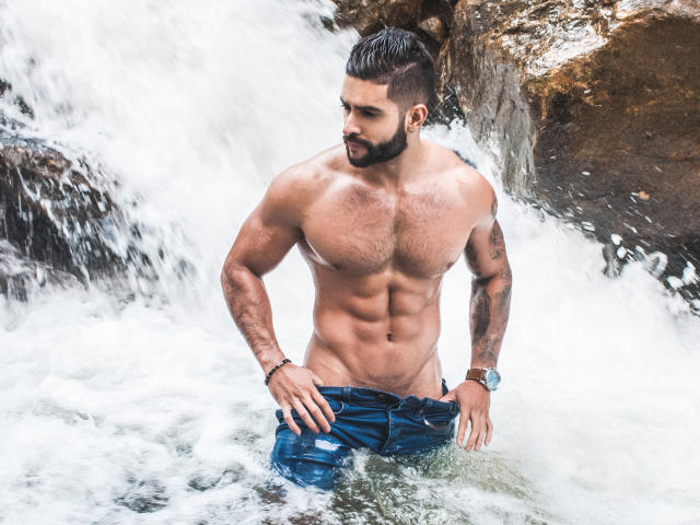 Lukas, shirtless with a muscular, hairy chest, stands in a waterfall, pulling down his blue jeans, showing a tattooed arm and a watch, looking to the side.