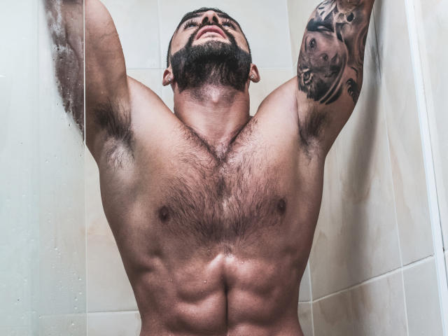 Lukas, muscular, hairy-chested, bearded man, arms raised, showering, tattoo on right arm, water on glass, visible abs, chest hair, and armpit hair, looking upward.