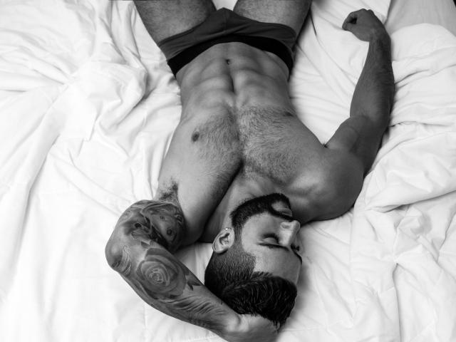 Lukas, muscular and hairy, lies on a bed with arms raised, wearing dark briefs, tattooed arm, and beard, with a strong, defined chest and abs in a black-and-white photo.