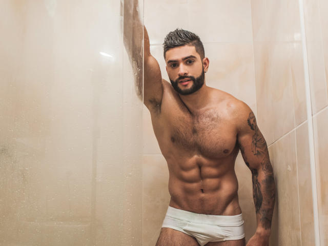 Muscular Lukas with dark hair and beard, wearing white underwear, leans against a glass shower wall, showing his hairy chest, tattoos, and defined abs. His arm is raised.
