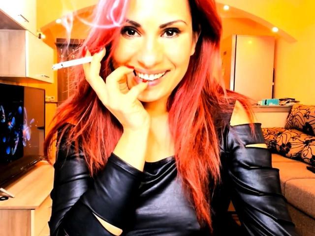 FantasyMe con cabello rojo, sonriendo, sosteniendo un cigarrillo en sus labios, vistiendo una chaqueta de cuero negra, en una habitación iluminada con luz amarilla, con un televisor a la izquierda..