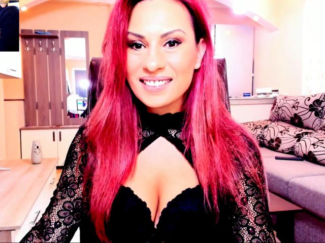 FantasyMe tiene el cabello rojo brillante, una blusa de encaje negro con un corte que deja ver su escote, mostrando sus senos, está sonriendo y está de pie en una habitación con un sofá gris y muebles de madera..