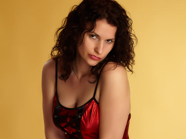 KinkyKosmina with curly dark brown hair, wearing a red satin lingerie top with black straps, showing her tits, and giving a slight, sexy smile against a yellow background.