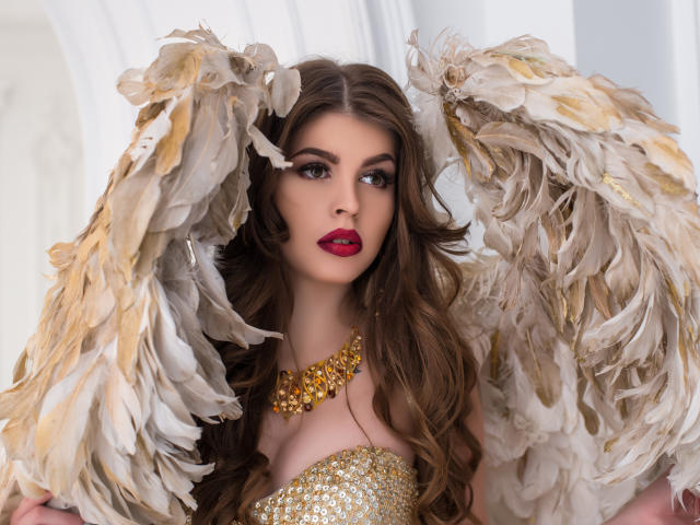 ValeriMair with big, feathery wings, golden necklace, and red lipstick, showing off her tits, with wavy brown hair and a sexy, confident expression.