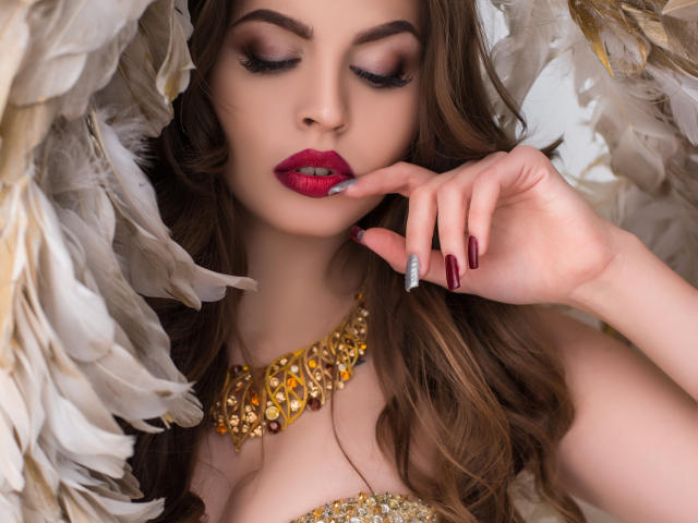 ValeriMair met rode lippenstift, bruin golvend haar, een gouden ketting en donkerrode nagels, die haar lip aanraakt, omringd door witte veren. Haar borsten zijn gedeeltelijk zichtbaar..