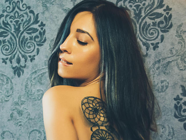 PoxyVibe with long dark hair, closed eyes, glossy lips, and a black floral tattoo on her back, showing off her tits. She's looking over her shoulder, smiling subtly.