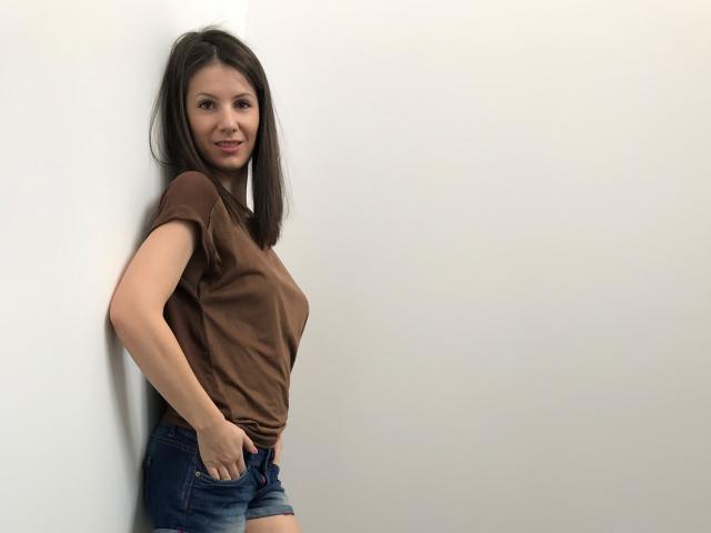 LanyJolene vestindo uma camiseta marrom, shorts de jeans azuis, com a mão no bolso, em pé contra uma parede branca, sorrindo, seios médios, cabelo escuro, pele clara, pose casual..