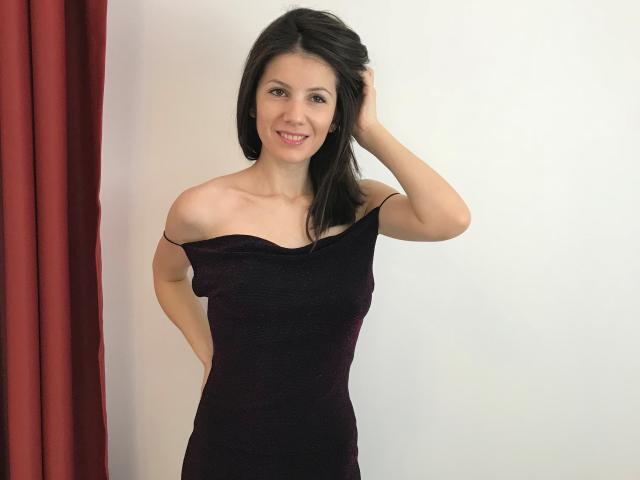 Lany Jolene mit schwarzen Haaren, lächelnd, trägt ein dunkelviolettes Kleid mit einem Schulterfrei-Schnitt, eine Hand im Haar, steht, roter Vorhang links, weiße Wand, zeigt ihre kleinen Brüste und ihren Hintern..