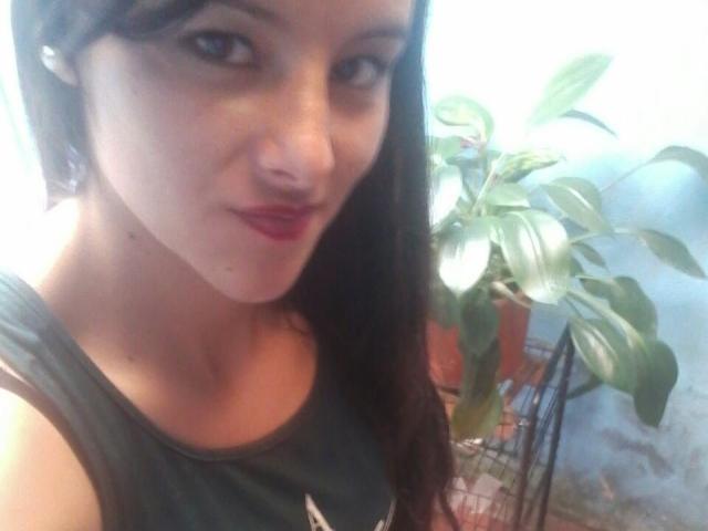 MiahottXX heeft een lichte huid, donkerbruin haar en rode lippenstift. Ze draagt een groene tanktop, waardoor haar kleine borsten zichtbaar zijn. Ze glimlacht licht en er staat een plant op de achtergrond..