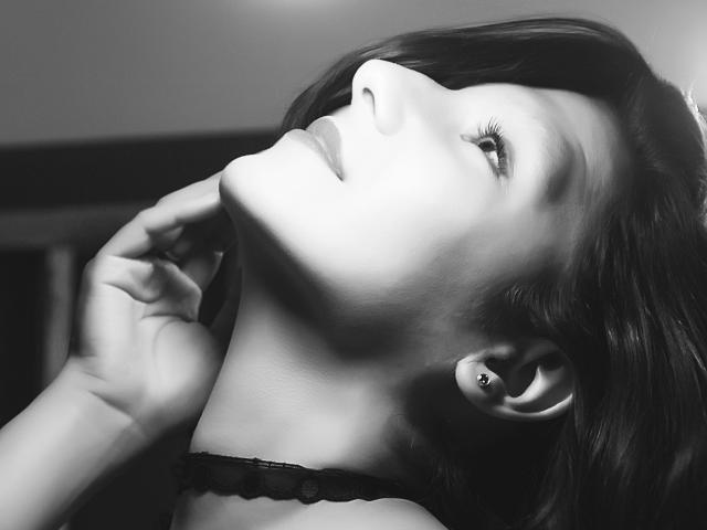 Black-and-white photo of AmethystCharm with chin raised, looking up, hand near neck, short dark hair, ear with small hoop earring, choker, soft light, slightly open mouth.