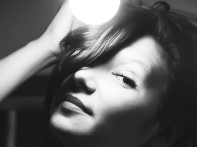 AmethystCharm's face is in black-and-white, lit by a bright light source above, with her eyes slightly open and lips gently parted, her hand touching her head.