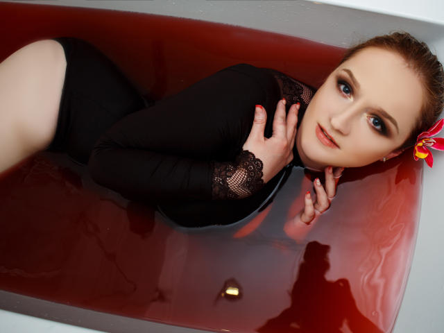 OhMollyHoney, met bleke huid, blauwe ogen en rood haar, draagt een zwart kanten body en ligt in badwater dat rood is gekleurd. Haar hand rust op haar borst, en er zit een bloem in haar haar..