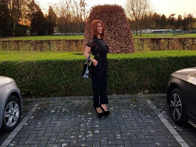 IssaDiamondd steht auf einem Parkplatz, trägt ein schwarzes Spitzenoberteil, schwarze Hosen und schwarze High Heels, hat rotes Haar, hält eine schwarze Tasche und präsentiert ihr Hinterteil..