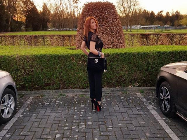 IssaDiamondd è in un parcheggio, indossa una camicetta di pizzo nera, pantaloni neri e scarpe con il tacco rosso, tiene una borsa nera, e ha un'auto da entrambi i lati..