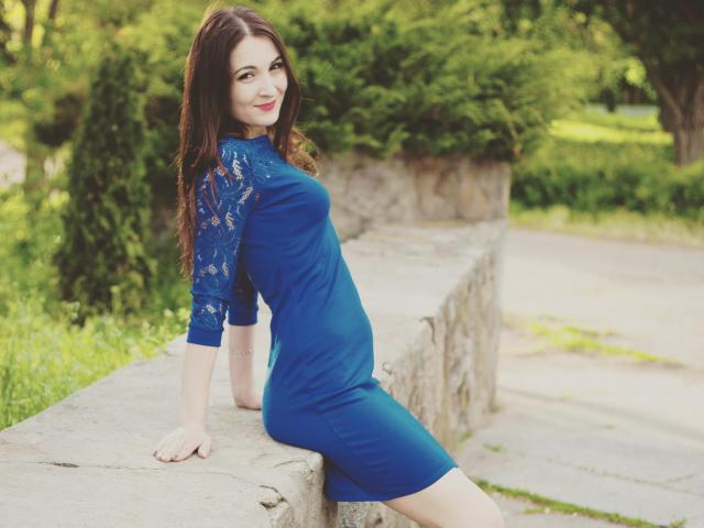Eleonorcka, con lunghi capelli castani, un abito di pizzo blu e rossetto rosso, seduta su un muro di pietra, sorride alla fotocamera, mostrando il sedere nell'abito..