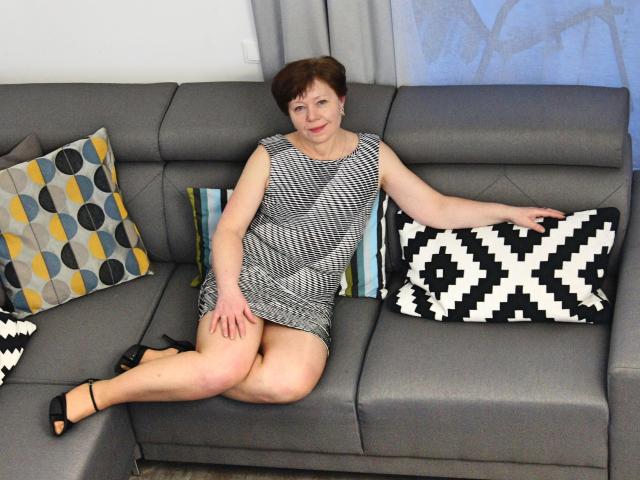 AdeleLoveEx, a short-haired woman with fair skin and red lipstick, wearing a black-and-white houndstooth dress, sits on a gray sofa with legs crossed, black heels on.