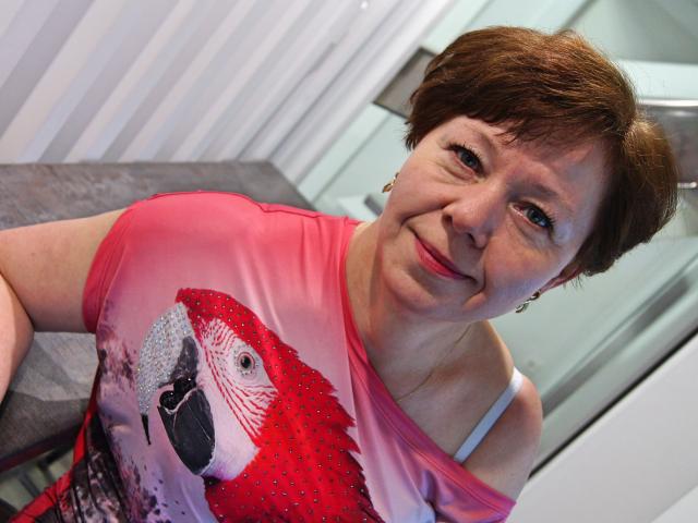 AdeleLoveEx, middle-aged woman with short brown hair, wearing a pink shirt with a red bird graphic, smiling slightly, showing a bit of a white bra strap.