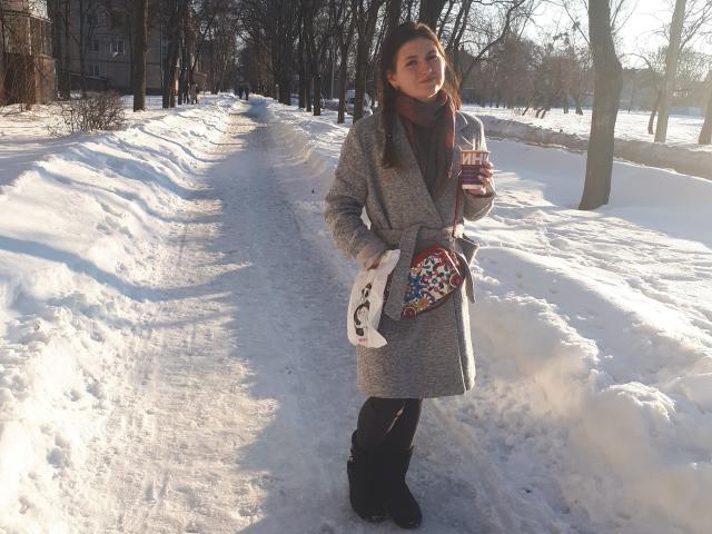 ShyNaomi è in piedi nella neve, con un cappotto grigio, tiene una tazza, indossa stivali neri e una borsa colorata, con una sciarpa bianca e capelli castani, con la luce del sole sul viso..