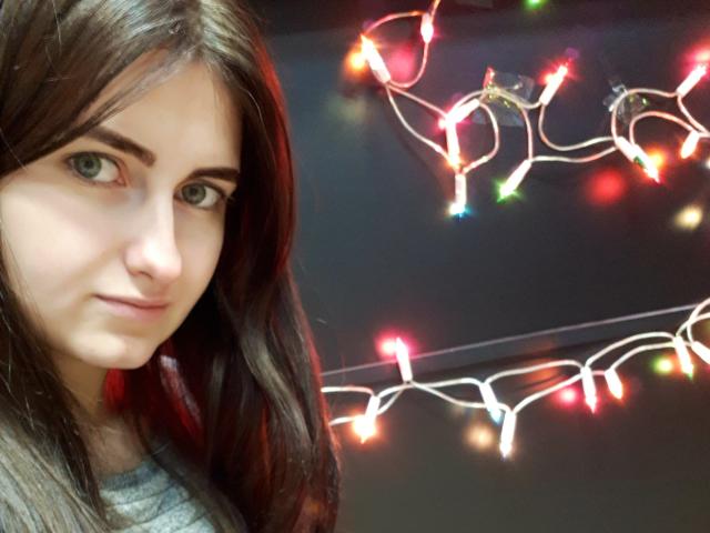 ShyNaomi, avec ses longs cheveux bruns, ses yeux verts et un léger sourire, porte une chemise grise. Des guirlandes de Noël colorées sont visibles en arrière-plan. Ses seins ne sont pas visibles..