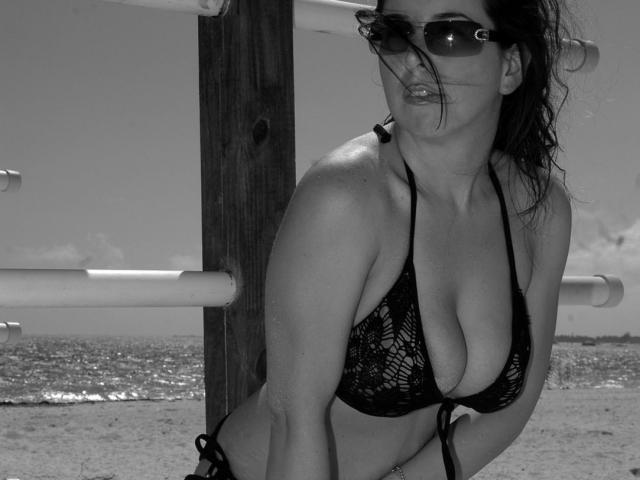 Black-and-white photo of TitanicTits with dark, wavy hair, sunglasses, and a lace bikini top showing her large, exposed tits. She leans against a wooden structure on a beach.