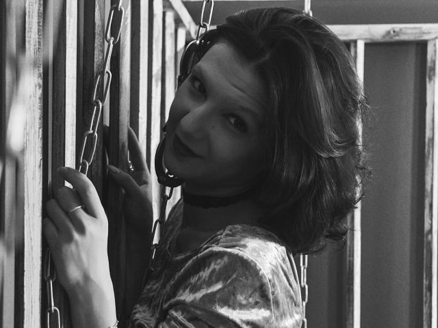 AmethystCharm with short, wavy hair, wearing a shiny, textured top, leaning against metal bars, looking over her shoulder, black choker, holding a chain, smiling subtly, black and white.